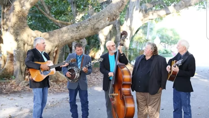 five older men in suit coats and jeans standing in front of a banyan tree holding a guitar, a banjo, an upright bass and another guitar.