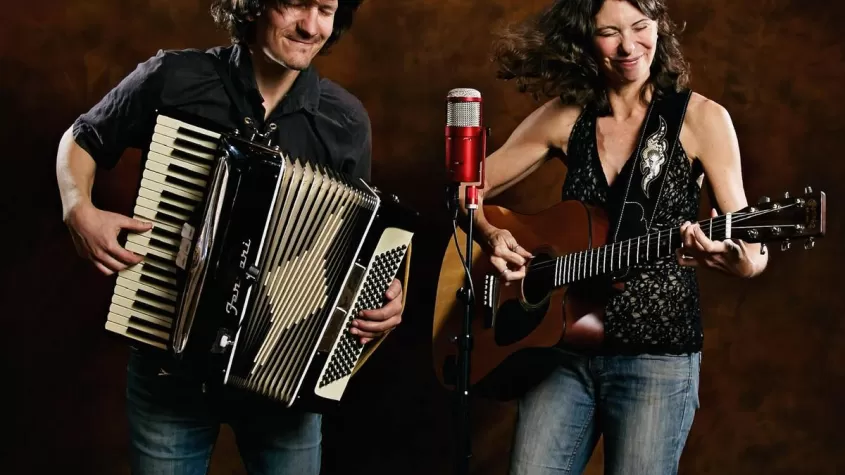 A man with long hair holding a piano accordion and a woman in a tank top holding a guitar in front of a red backdrop with a red microphone