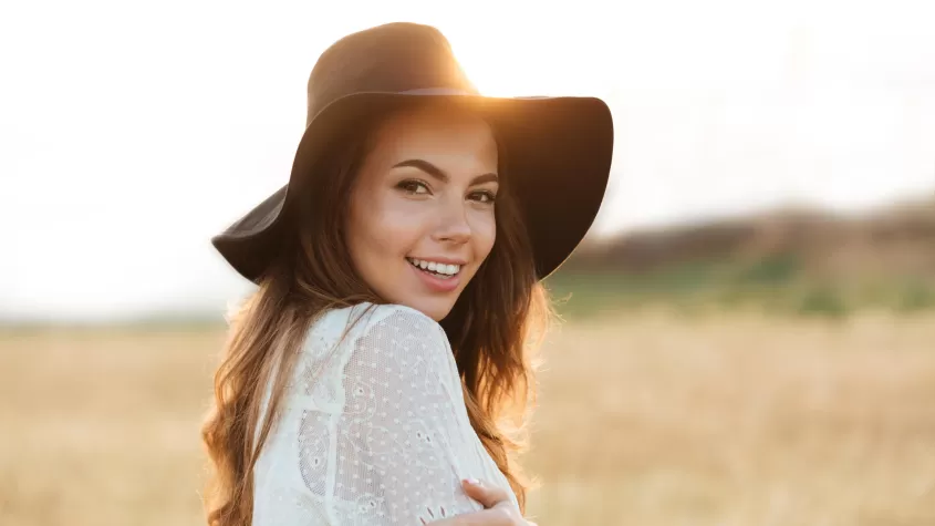 image of a woman smiling in a hay field with a hat on and the sun behind her