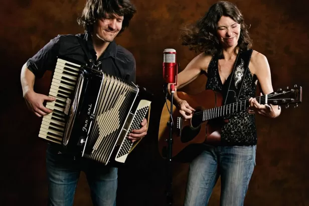 A man with long hair holding a piano accordion and a woman in a tank top holding a guitar in front of a red backdrop with a red microphone
