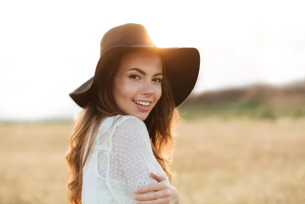 image of a woman smiling in a hay field with a hat on and the sun behind her
